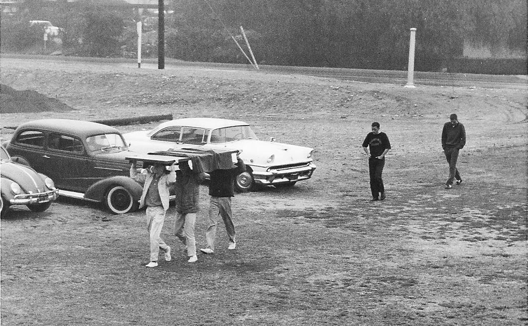 3 students carry a door on their heads across a field, followed by 2 others. 