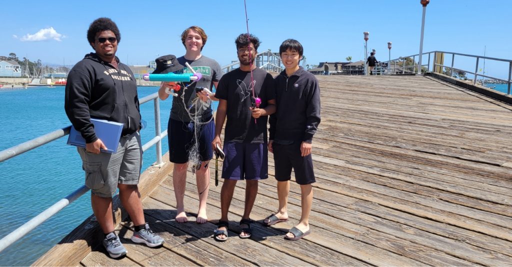 Four students stand on wooden pier; one hold underwater remove vehicle.