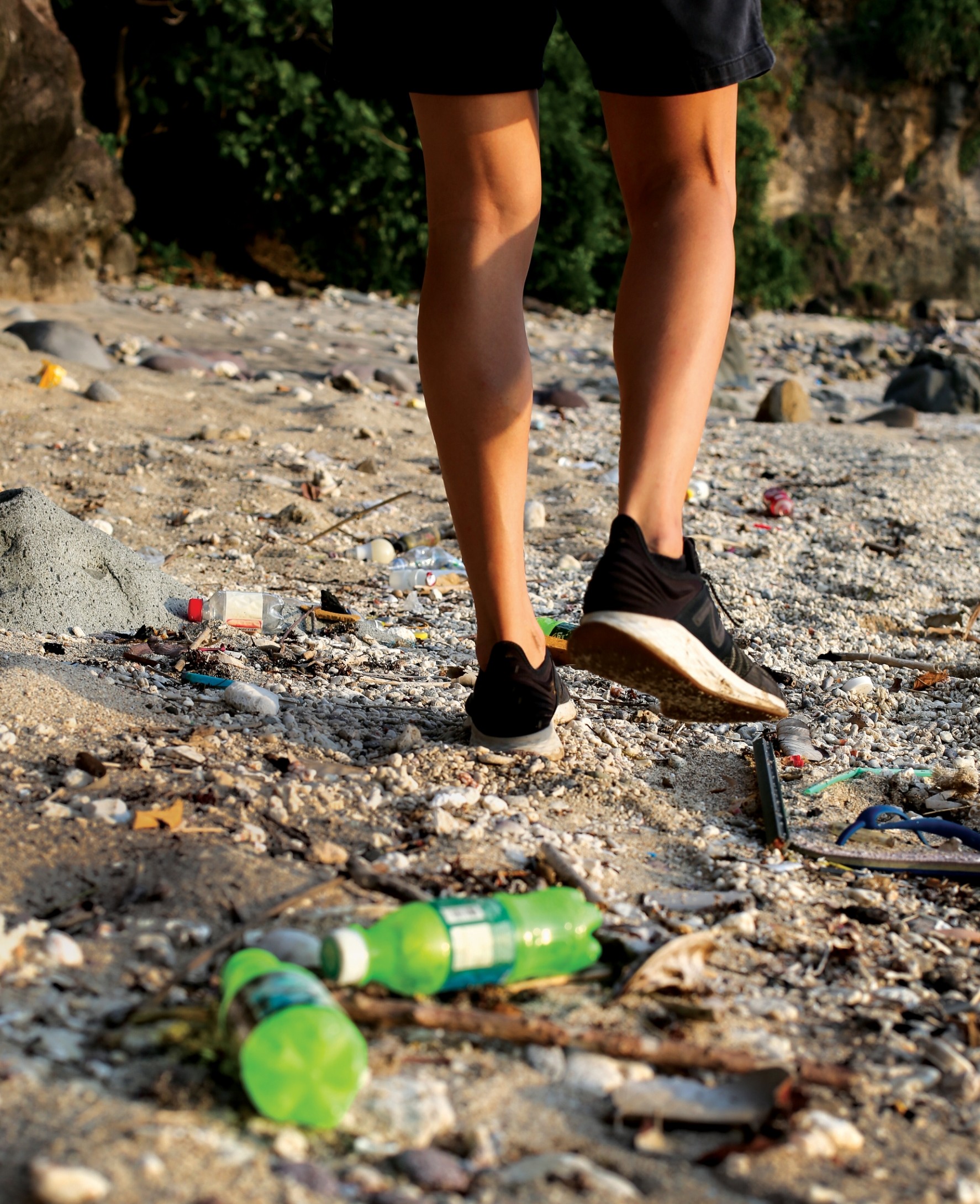 A person with only legs visible walks a trash-strewn beach.